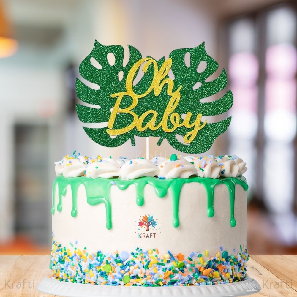Oh Baby cake topper with green glitter monstera leaf and gold glitter lettering placed on a frosted cake for a baby shower celebration